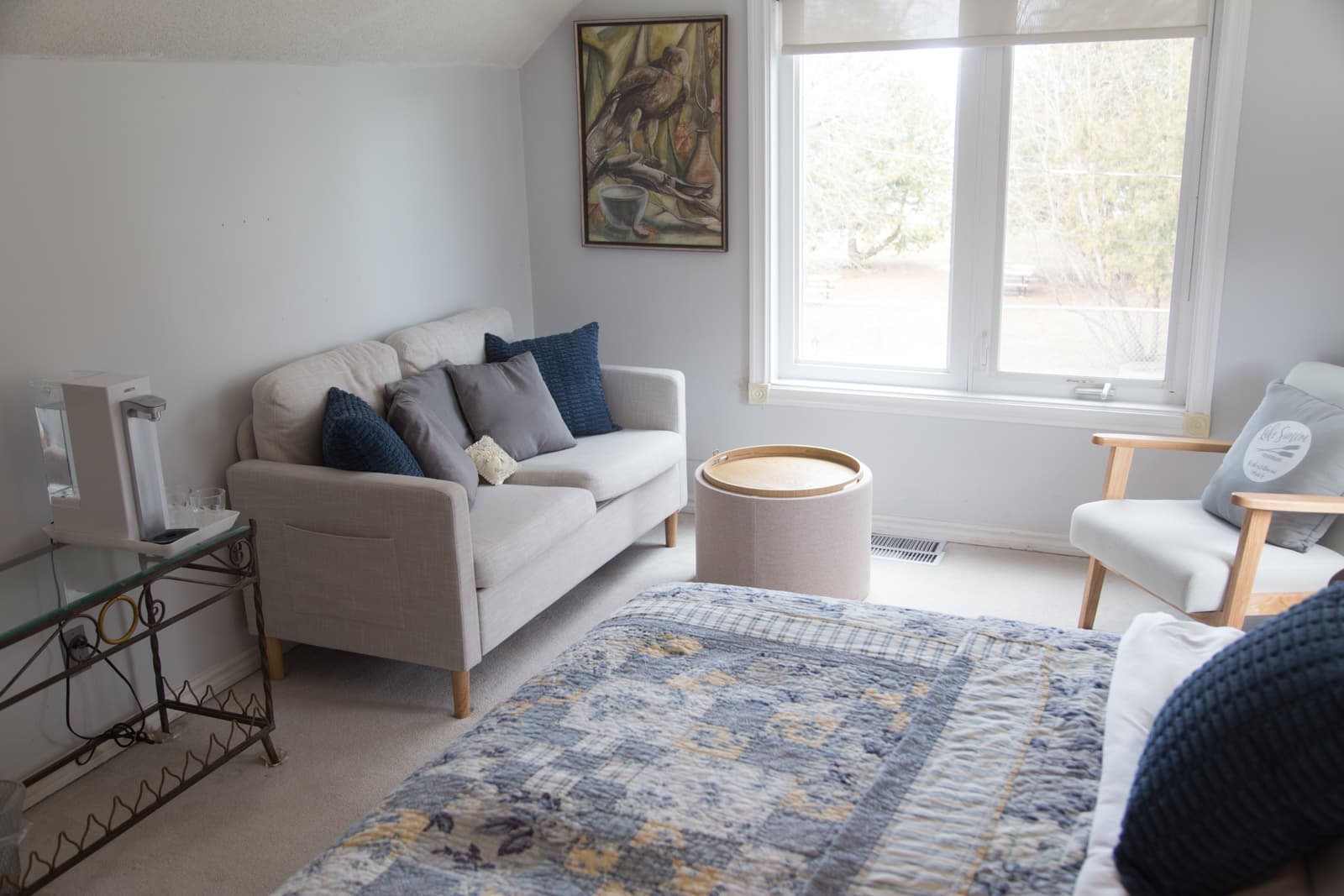 The sitting area of the Blue Room — a pale sofa with navy and grey cushions, a round ottoman and a wooden-armed chair by the window